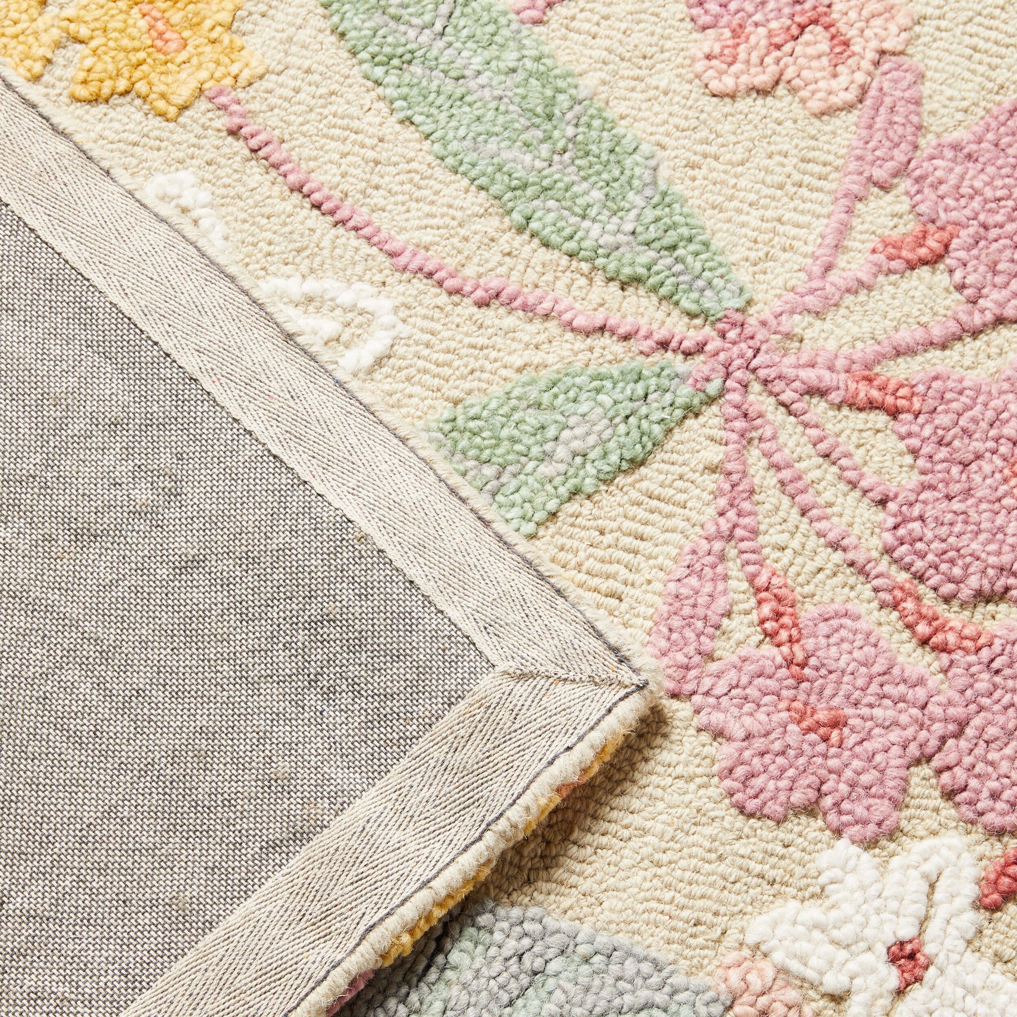 A corner of the Botanic Native Rug 240x150cm showcases its beige-bordered floral design in pink, green, and yellow tones. The hand-tufted rug, made from 100% wool, is partially flipped to show the gray underside.