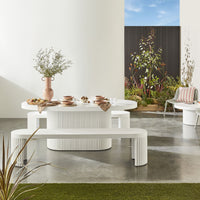 Transform your concrete patio into a stylish haven with the modern elegance of our Ripple Curved Outdoor Dining Table 200cm and Bench Package, featuring sleek white ribbed furniture, lush greenery, and contemporary tableware.